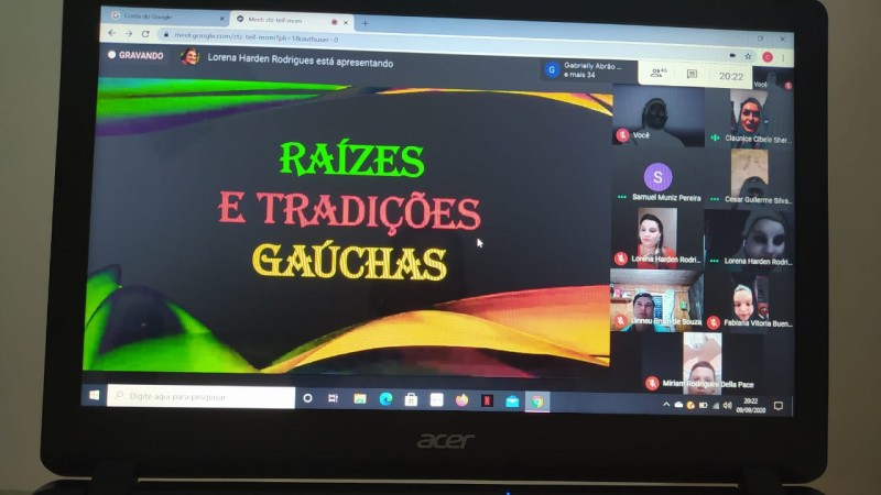 Foto registrada durante a videoconferência, na qual aparecem pessoas assistindo à palestra pelo Google Meet e um cartaz escrito "Raízes e tradições gaúchas"