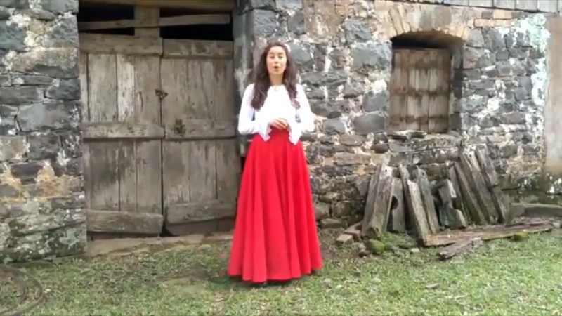 Print screen tirado do vídeo feito pelos alunos da Escola Pedro Migliorini, onde aparece uma aluna, vestida de prenda, em frente a um galpão feito de pedra.