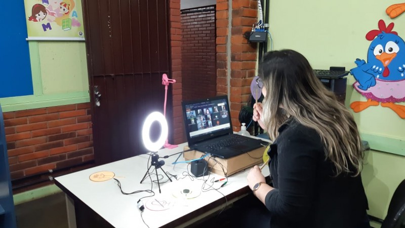 Foto de uma professora de costas, sentada e utilizando o estúdio, no qual equipamentos como notebook e ring light