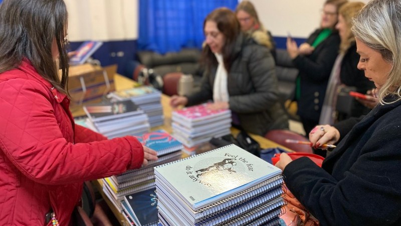Foto tirada na sala dos professores da Escola Eldorado do Sul. Em primeiro plano, estão uma série de cadernos escolares, colocados em cima de uma mesa de madeira. A equipe diretiva da escola está ao redor da mesa, organizando os cadernos para distribuir aos alunos.