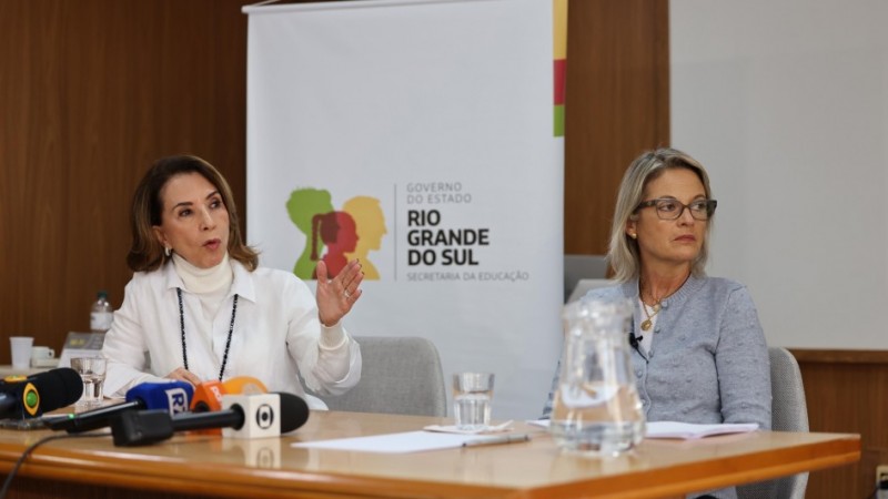 Na imagem, há duas mulheres sentadas à mesa em uma coletiva de imprensa. À esquerda, está Raquel Teixeira, a secretária de Educação do Rio Grande do Sul. Ela tem cabelo castanho claro, liso e na altura dos ombros, e está usando uma blusa branca de gola alta com um colar comprido de contas escuras. Ela gesticula com a mão direita enquanto fala. À direita, está Izabel Matte, a secretária de Obras Públicas. Ela tem cabelo loiro, liso e também na altura dos ombros, e usa óculos de armação preta. Izabel veste um cardigã cinza sobre uma blusa clara e está olhando para a frente com uma expressão séria. No fundo, há um banner com o logotipo do Governo do Estado do Rio Grande do Sul e o texto "Secretaria da Educação". Sobre a mesa, há diversos microfones de diferentes veículos de comunicação, um jarro de vidro com água e alguns papéis.