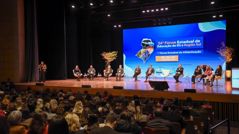 A imagem mostra o auditório da Feevale. No palco, há várias pessoas sentadas em cadeiras, formando um painel de discussão. A secretária adjunta Stefanie está mais à esquerda, de pé, falando ao microfone, enquanto os demais membros do painel estão sentados. O fundo do palco exibe uma grande tela com o texto "34º Fórum Estadual de Educação do RS e Região Sul" e "1º Fórum Estadual de Alfabetização", seguido das datas "03, 04 e 05 de setembro 2024". A tela também mostra mapas da região e o logotipo da Undime-RS (União dos Dirigentes Municipais de Educação do Rio Grande do Sul). A plateia, composta por muitas pessoas, está sentada assistindo à discussão