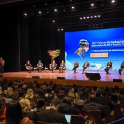 A imagem mostra o auditório da Feevale. No palco, há várias pessoas sentadas em cadeiras, formando um painel de discussão. A secretária adjunta Stefanie está mais à esquerda, de pé, falando ao microfone, enquanto os demais membros do painel estão sentados. O fundo do palco exibe uma grande tela com o texto "34º Fórum Estadual de Educação do RS e Região Sul" e "1º Fórum Estadual de Alfabetização", seguido das datas "03, 04 e 05 de setembro 2024". A tela também mostra mapas da região e o logotipo da Undime-RS (União dos Dirigentes Municipais de Educação do Rio Grande do Sul). A plateia, composta por muitas pessoas, está sentada assistindo à discussão