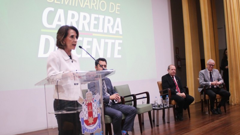 Raquel em pé, atrás de um púlpito e falando ao microfone sobre o placo no Seminário de Carreira Docente 2024. Ela é observada por quatro homens que compõem a mesa de abertura do evento.