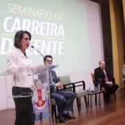 Raquel em pé, atrás de um púlpito e falando ao microfone sobre o placo no Seminário de Carreira Docente 2024. Ela é observada por quatro homens que compõem a mesa de abertura do evento.