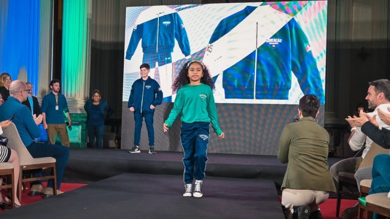 Uma menina caminha em uma passarela vestindo o novo uniforme escolar da rede estadual, composto por calça azul e camiseta verde. Ao fundo, um garoto também exibe o uniforme diante de um telão que mostra detalhes das peças. O público ao redor aplaude enquanto assiste ao desfile.