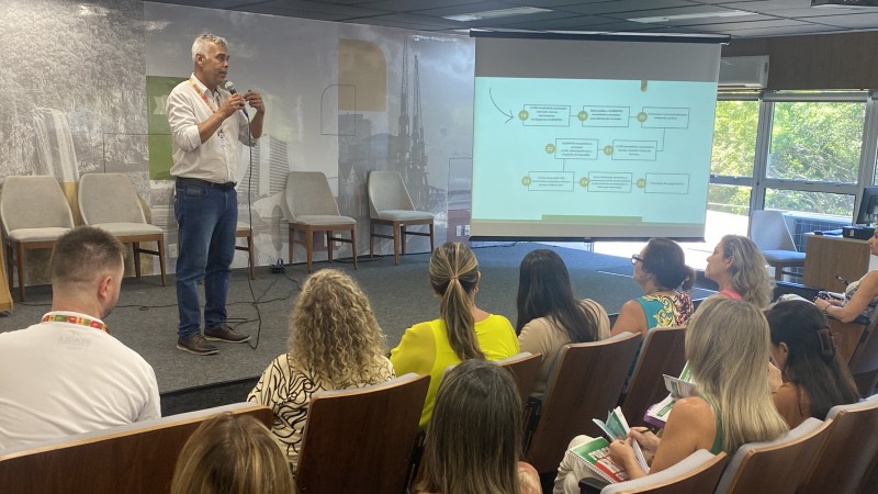 A imagem mostra o subsecretário Rômulo Campos, um homem de camisa branca e jeans, falando ao microfone em um auditório do Centro Administrativo Fernando Ferrari, em Porto Alegre. Ele está ao lado de uma tela projetada com um fluxograma. A plateia segura materiais impressos enquanto acompanha a apresentação.







