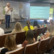 A imagem mostra o subsecretário Rômulo Campos, um homem de camisa branca e jeans, falando ao microfone em um auditório do Centro Administrativo Fernando Ferrari, em Porto Alegre. Ele está ao lado de uma tela projetada com um fluxograma. A plateia segura materiais impressos enquanto acompanha a apresentação.







