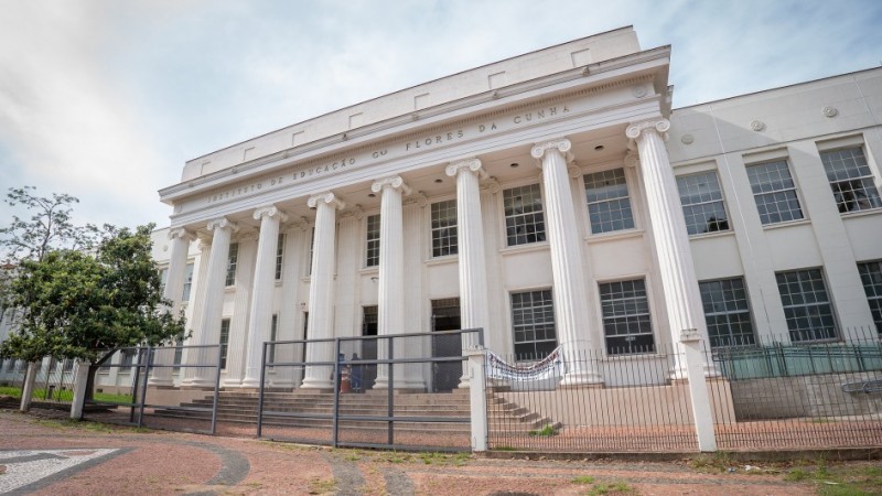 Fachada do Instituto Estadual Flores da Cunha, em Porto Alegre, um prédio de arquitetura neoclássica. A construção possui colunas brancas imponentes e diversas janelas simétricas. Há uma grade ao redor da entrada principal e uma faixa pendurada no portão.