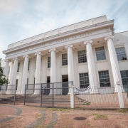 Fachada do Instituto Estadual Flores da Cunha, em Porto Alegre, um prédio de arquitetura neoclássica. A construção possui colunas brancas imponentes e diversas janelas simétricas. Há uma grade ao redor da entrada principal e uma faixa pendurada no portão.