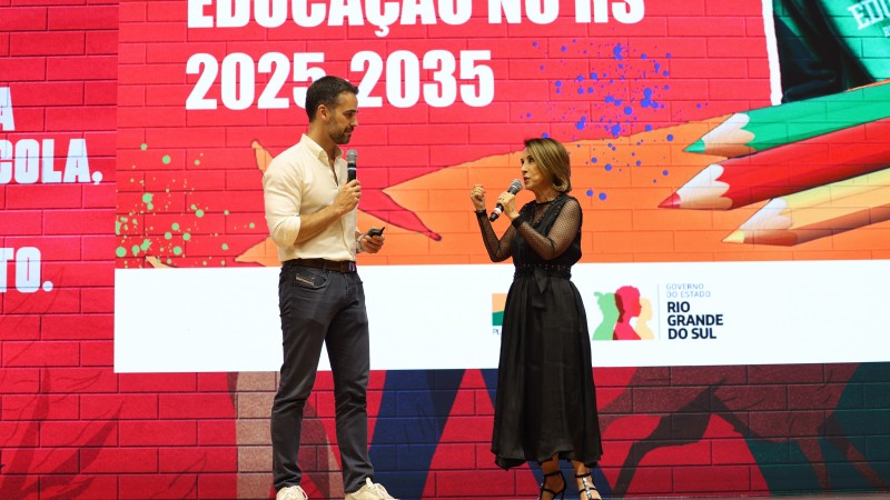 A foto mostra Leite e a secretária da Educação Raquel Teixeira no palco da casa da Ospa, em Porto Alegre. Ao fundo, há um grande painel vermelho com a inscrição "EDUCAÇÃO NO RS 2025-2035" e o logotipo do Governo do Estado do Rio Grande do Sul. O cenário tem elementos gráficos coloridos relacionados à educação, como lápis e formas abstratas.