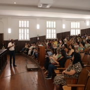 Foto do auditório do Instituto de Educação, em Porto Alegre, onde uma plateia de professores escuta o subsecretário de desenvolvimento da educação, Marcelo Jerônimo, que segura um microfone próximo a uma mesa de madeira. 