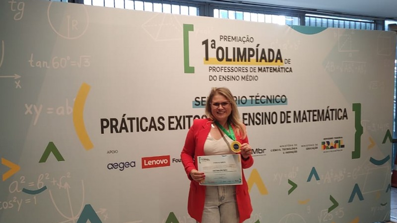 Foto de Joséli Pagliosa, uma mulher loira de óculos. Na imagem, ela está vestindo blazer vermelho e calça jeans, segura um certificado e usa uma medalha no pescoço. Ela está diante de um painel com fórmulas matemáticas e o texto "1ª Olimpíada de Professores de Matemática do Ensino Médio". O ambiente tem piso de mármore e iluminação moderna.