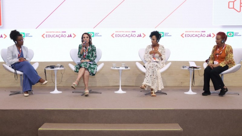 Raquel participando de um debate com mais três mulheres negras no Encontro Anual Educação Já 2025. Elas estão sentadas em cadeiras e um palco. Raquel fala ao microfone.