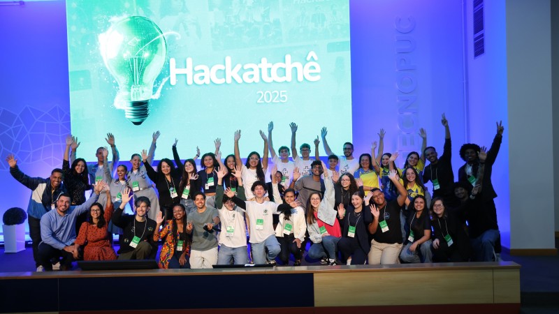 Grupo diverso de estudantes e educadores posa sorridente com braços erguidos em frente a um telão com a logo “Hackatchê 2025”. A imagem foi registrada em um auditório moderno com iluminação azul e a marca “Tecnopuc” projetada na parede lateral.