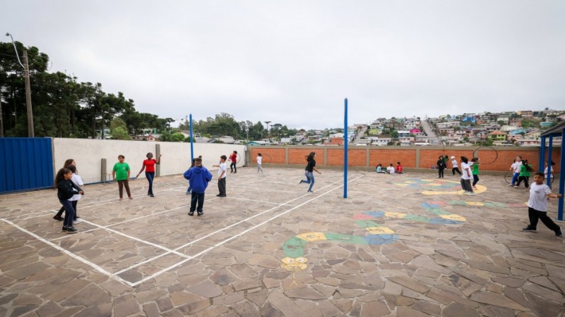 Foto de pátio escolar com dezenas de crianças brincando. O muro em volta é de tijolo à vista, parede branca e com um portão na cor azul. No chão há pinturas de desenhos coloridos e uma marcação de quadra de vôlei.