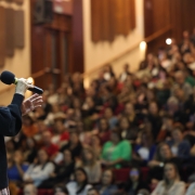 Foto da secretária da Educação, Raquel Teixeira, segurando um microfone preto, diante de uma plateia de diretores e orientadores que aparecem ao fundo sentados.