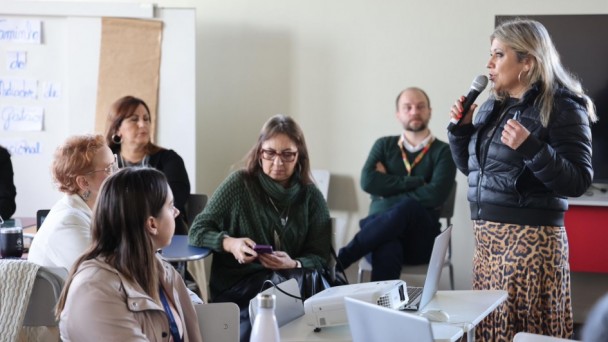 Subsecretária Néri, um mulher loira de altura mediana, está em pé, falando ao microfone diante de um grupo sentado em uma sala de aula. Ela veste casaco preto e saia estampada. Os participantes observam atentamente, com notebooks e projetor sobre a mesa
