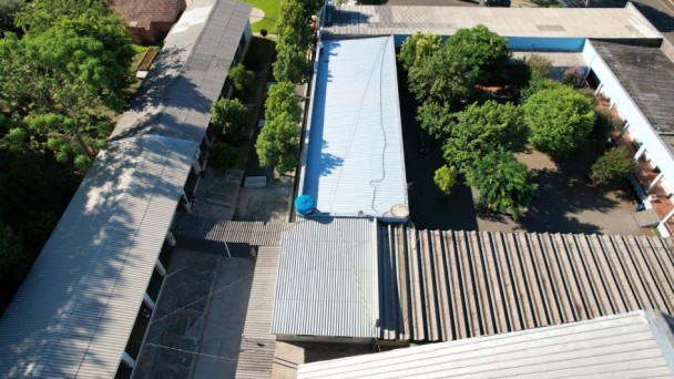 Imagem aérea da Escola Barão do Rio Branco, formada por um conjunto de edifícios com telhados metálicos, rodeado por árvores. Há áreas pavimentadas entre os prédios escolares e carros estacionados ao fundo, próximos a uma rua.