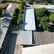 Imagem aérea da Escola Barão do Rio Branco, formada por um conjunto de edifícios com telhados metálicos, rodeado por árvores. Há áreas pavimentadas entre os prédios escolares e carros estacionados ao fundo, próximos a uma rua.