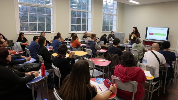 Sala de aula no Instituto de Educação General Flores da Cunha, em Porto Alegre, com pessoas sentadas em mesas coloridas, assistindo a uma apresentação feita por  uma mulher em pé, diante de uma tela grande. Ao fundo, há três janelas amplas.
