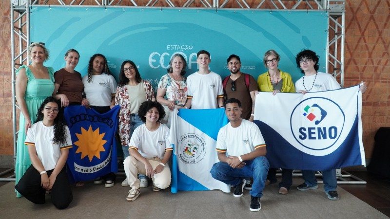 Grupo de estudantes e professores da Rede Estadual posa diante de um painel, segurando três bandeiras: uma com emblema de sol, outra azul e branca, e uma com a palavra "Seno", representando as escolas estaduais