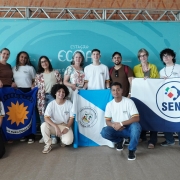 Grupo de estudantes e professores da Rede Estadual posa diante de um painel, segurando três bandeiras: uma com emblema de sol, outra azul e branca, e uma com a palavra "Seno", representando as escolas estaduais