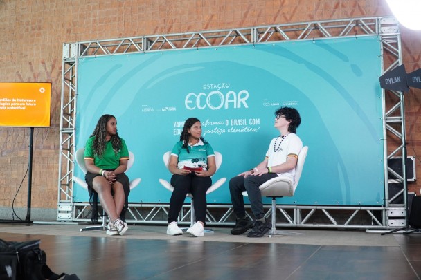 Três jovens participam de um painel no evento "Estação Ecoar". Eles estão sentados em cadeiras diante de um painel azul com o tema do evento ao fundo, discutindo educação e justiça climática.