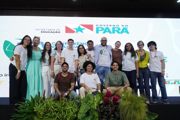 Grupo diverso de estudantes e educadores posa sorridente em frente a um painel com a marca do Governo do Pará e da Secretaria de Educação, durante evento com temática ambiental. Há plantas decorativas em primeiro plano.