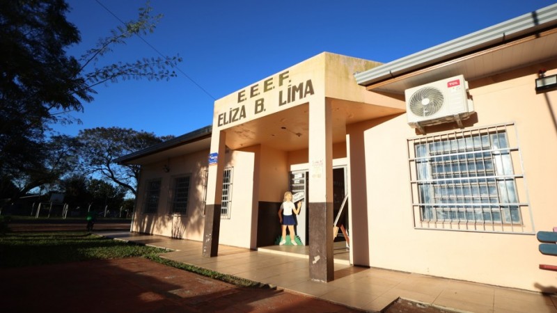 A imagem mostra a entrada principal de uma escola em um dia ensolarado. A fachada do pr&eacute;dio &eacute; bege e marrom escuro na parte inferior. Acima da porta de entrada, em letras grandes e escuras, l&ecirc;-se "E. E. E. F. ELIZA B. LIMA". &Agrave; esquerda da porta, h&aacute; um adesivo vermelho e azul com a palavra "ROTA" e a frase "Seguran&ccedil;a em Primeiro Lugar". No canto superior direito da imagem, um aparelho de ar condicionado est&aacute; fixado na parede externa. Atrav&eacute;s da porta aberta, &eacute; poss&iacute;vel ver um interior iluminado e algumas figuras de papel&atilde;o, incluindo uma representa&ccedil;&atilde;o de uma menina e um cavalete com um desenho colorido.