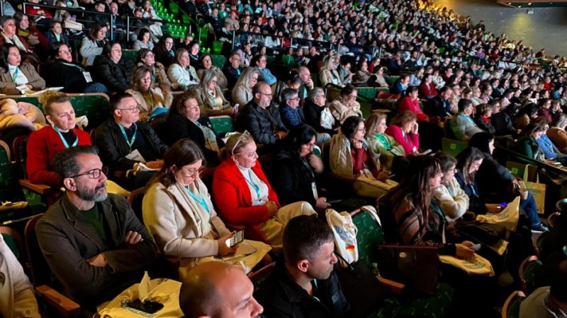 Audit&oacute;rio lotado com centenas de pessoas sentadas, todas voltadas para o palco, assistindo atentamente a uma apresenta&ccedil;&atilde;o. A ilumina&ccedil;&atilde;o foca no p&uacute;blico da frente, e muitos usam crach&aacute;s, indicando participa&ccedil;&atilde;o no Semin&aacute;rio da Secretaria da Educa&ccedil;&atilde;o.