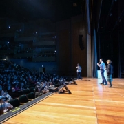 Auditório lotado com várias pessoas assistindo a uma apresentação. Três pessoas estão no palco de madeira, uma fala ao microfone. Plateia atenta, alguns registros com celular. Ambiente amplo e com vários níveis de assentos.