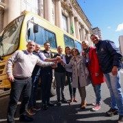 Esta é uma fotografia de um grupo de oito pessoas, seis homens e duas mulheres, em pé na rua, posando para a câmera em frente a um grande ônibus escolar amarelo. O grupo está disposto em um semicírculo. Todos sorriem e estendem as mãos em direção ao centro, como em um aperto de mão coletivo. O homem no centro, com barba, veste um terno azul-marinho. A mulher à sua direita veste um casaco bege de estilo trench coat. O ônibus amarelo, com a marca "IVECO", domina o lado esquerdo da imagem. Ao fundo, há um grande prédio com arquitetura formal, colunas e janelas altas. O céu é azul com nuvens brancas. O ambiente é de celebração e oficial.