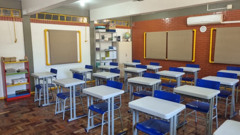Sala de aula de uma escola estadual do Rio Grande do Sul, organizada com carteiras e cadeiras azuis dispostas em fileiras. Ao fundo, h&aacute; estantes com livros, um arm&aacute;rio branco e um globo. Nas paredes, quadros de avisos com moldura amarela e um rel&oacute;gio pr&oacute;ximo ao ar-condicionado.