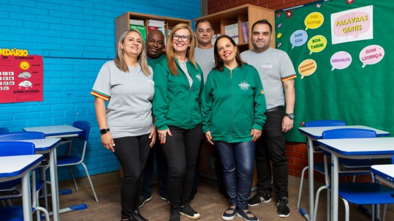 Seis pessoas adultas posam sorridentes dentro de uma sala de aula colorida, com paredes azuis e vermelhas. Tr&ecirc;s vestem camisetas cinzas e tr&ecirc;s usam casacos verdes com o logotipo &ldquo;Educa&ccedil;&atilde;o &ndash; Rede Estadual&rdquo; e a bandeira do Rio Grande do Sul na manga. Ao fundo, h&aacute; carteiras escolares azuis, cartazes educativos e uma estante com livros.