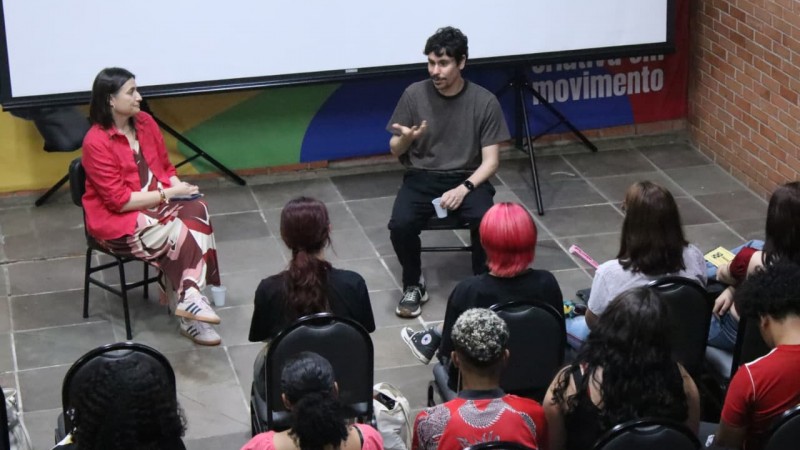 Pessoa mediadora e cineasta convidado conversam diante de um grupo de estudantes sentados. A mediadora est&aacute; &agrave; esquerda, enquanto o convidado est&aacute; ao centro, gesticulando. O p&uacute;blico observa a fala atentamente em uma sala com cadeiras e projetor ao fundo.