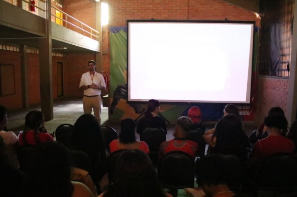 O superintendente Tomás Collier, um homem alto com roupas brancas, apresenta-se diante de um grupo de estudantes sentados, ao lado de uma tela de projeção iluminada. A sala é ampla, com paredes de tijolos e luz natural entrando por cima. O público observa atentamente a fala de Collier.