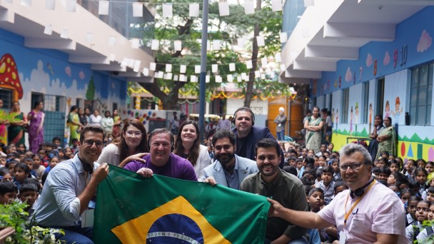 Grupo de autoridades da comitiva brasileira posa sorrindo com uma bandeira do Brasil em uma escola da &iacute;ndia, decorada com murais coloridos. Atr&aacute;s deles, muitas crian&ccedil;as sentadas observam a cena, enquanto adultos acompanham ao fundo no p&aacute;tio arborizado.