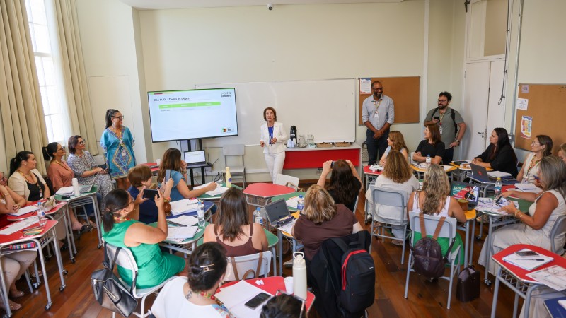 Sala de aula com cerca de 20 educadores sentados em mesas organizadas em c&iacute;rculo. Ao centro, a secret&aacute;ria da Educa&ccedil;&atilde;o, Raquel Teixeira, veste um terno branco enquanto fala ao grupo. H&aacute; um tel&atilde;o com apresenta&ccedil;&atilde;o, cadernos, notebooks e garrafas de &aacute;gua. Pessoas observam e fazem anota&ccedil;&otilde;es.