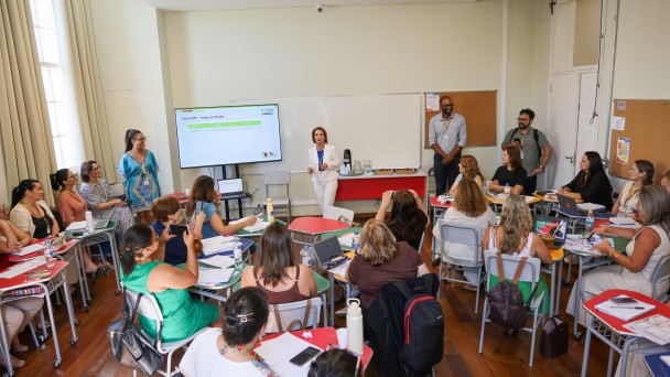 Sala de aula com cerca de 20 educadores sentados em mesas organizadas em c&iacute;rculo. Ao centro, a secret&aacute;ria da Educa&ccedil;&atilde;o, Raquel Teixeira, veste um terno branco enquanto fala ao grupo. H&aacute; um tel&atilde;o com apresenta&ccedil;&atilde;o, cadernos, notebooks e garrafas de &aacute;gua. Pessoas observam e fazem anota&ccedil;&otilde;es.