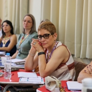 Mulheres sentadas em sala de aula participam de uma formação. Em primeiro plano, uma mulher de óculos apoia o rosto nas mãos e observa atentamente. Sobre as mesas há cadernos, canetas, garrafas de água e materiais de apoio. Ao fundo, outros participantes acompanham a atividade.