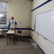 Homem de costas trabalha na parede de uma sala de aula com paredes, piso, pintura e móveis desgastados