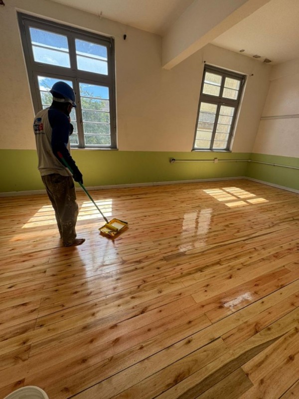 Homem pinta piso de sala com duas grandes janelas. Piso está muito brilhoso.

