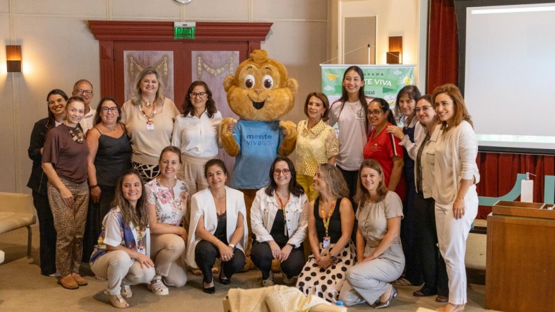 Grupo de pessoas posa para foto em ambiente interno de evento. Ao centro, um mascote sorridente com camiseta &ldquo;Mente Viva&rdquo;. Participantes est&atilde;o em p&eacute; e agachados, pr&oacute;ximos a um tel&atilde;o e banner do programa, em clima de integra&ccedil;&atilde;o.