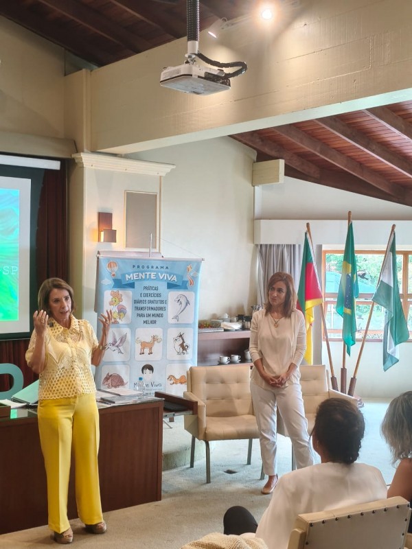 Duas mulheres palestram em sala com bandeiras ao fundo. À esquerda, uma mulher de amarelo gesticula ao falar ao lado de um banner do "Programa Mente Viva". À direita, outra mulher em tons claros observa. O ambiente possui teto de madeira e parte da plateia é visível em primeiro plano.