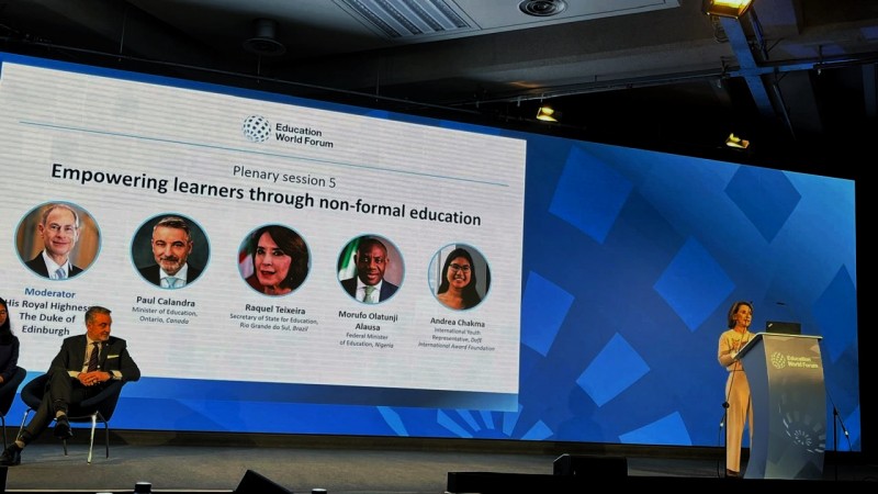 Audit&oacute;rio de confer&ecirc;ncia com painel do Education World Forum exibindo sess&atilde;o sobre educa&ccedil;&atilde;o n&atilde;o formal. &Agrave; direita, a secret&aacute;ria da Educa&ccedil;&atilde;o, Raquel Teixeira, fala em p&uacute;lpito no palco; &agrave; esquerda, participantes est&atilde;o sentados. A tela mostra nomes e fotos dos debatedores internacionais.
