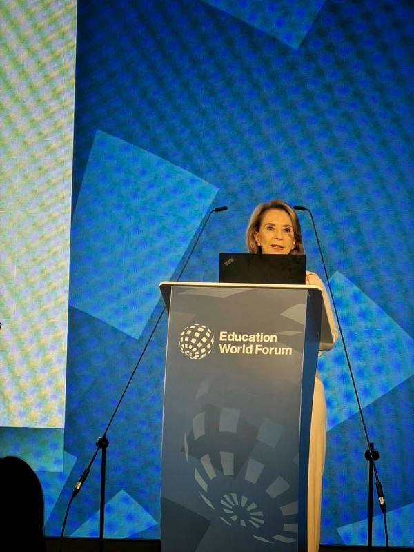 A secretária da Educação, Raquel Teixeira, aparece em pé atrás de um púlpito no palco do Education World Forum, diante de um fundo azul com formas geométricas. Ela fala ao microfone enquanto utiliza um notebook apoiado no púlpito.