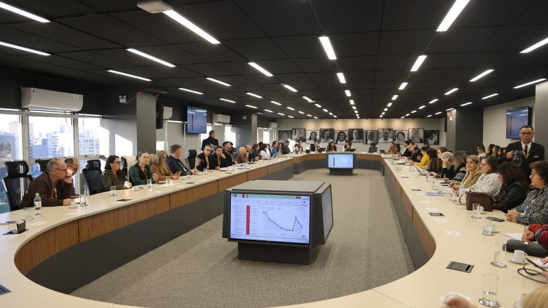 Fotografia de uma ampla sala de reuni&otilde;es com dezenas de pessoas sentadas ao redor de uma grande mesa em formato de 'U'. No centro, monitores exibem gr&aacute;ficos. O ambiente possui teto escuro com ilumina&ccedil;&atilde;o linear, grandes janelas &agrave; esquerda e retratos em preto e branco na parede ao fundo.