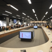 Fotografia de uma ampla sala de reuni&otilde;es com dezenas de pessoas sentadas ao redor de uma grande mesa em formato de 'U'. No centro, monitores exibem gr&aacute;ficos. O ambiente possui teto escuro com ilumina&ccedil;&atilde;o linear, grandes janelas &agrave; esquerda e retratos em preto e branco na parede ao fundo.