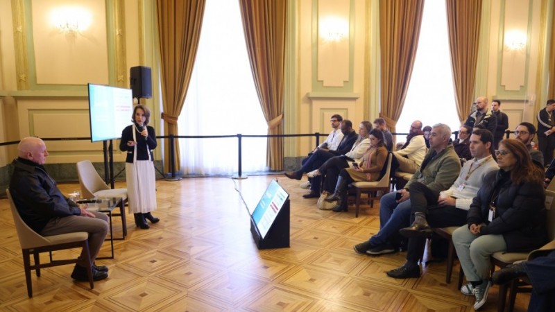 Sala ampla com piso de madeira e cortinas altas. A secret&aacute;ria da Educa&ccedil;&atilde;o, Raquel Teixiera, apresenta uma palestra com microfone ao lado de uma tela, enquanto um o secret&aacute;rio de Comunica&ccedil;&atilde;o, Caio Tomazeli, est&aacute; sentado &agrave; frente. Cerca de 15 pessoas acompanham a apresenta&ccedil;&atilde;o, sentadas em fileiras. H&aacute; um monitor no ch&atilde;o exibindo os slides.